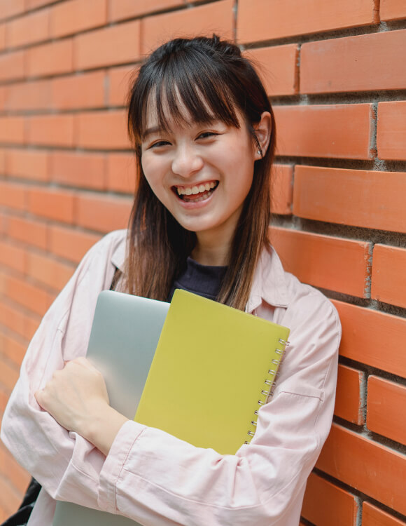 Smiling teenage female student holding school materials and leaning on a brick wall