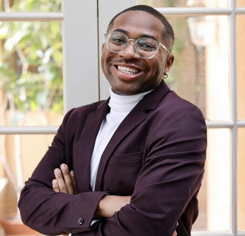 A man stands in front of a white window. He wears a white turtle neck and a maroon suit jacket and clear glasses. He has his arms crossed and smiles widely at the camera.