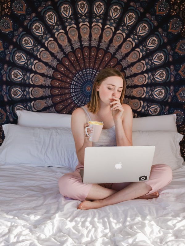 A girl sits on a bed cross legged. Her sheets are completely white and there is a tapestry on the wall behind her. She holds a cup of coffee and looks down at a laptop in her lap.