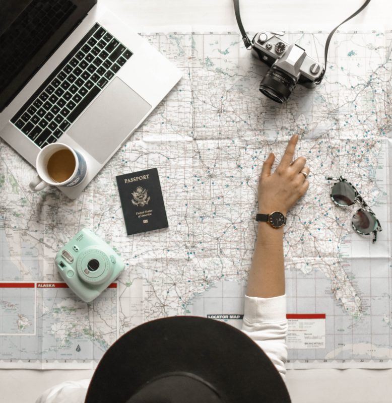 A white map of the United States lays on a table. The camera looks from above. A girl with a black hat looks over the map and points at Michigan. On the map sits a laptop, a coffee cup, a Polaroid camera, a passport, a camera, and a pair of sunglasses.