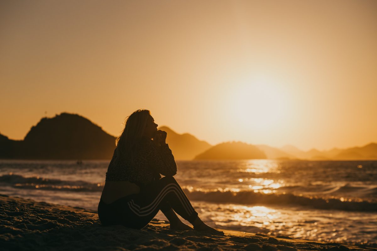 A girl sits on the beach next to the ocean. An orange sunset is in the sky behind her. Her head is rested on her hands and she stares pensively into the ocean. She wears a sweatshirt and adidas pants.