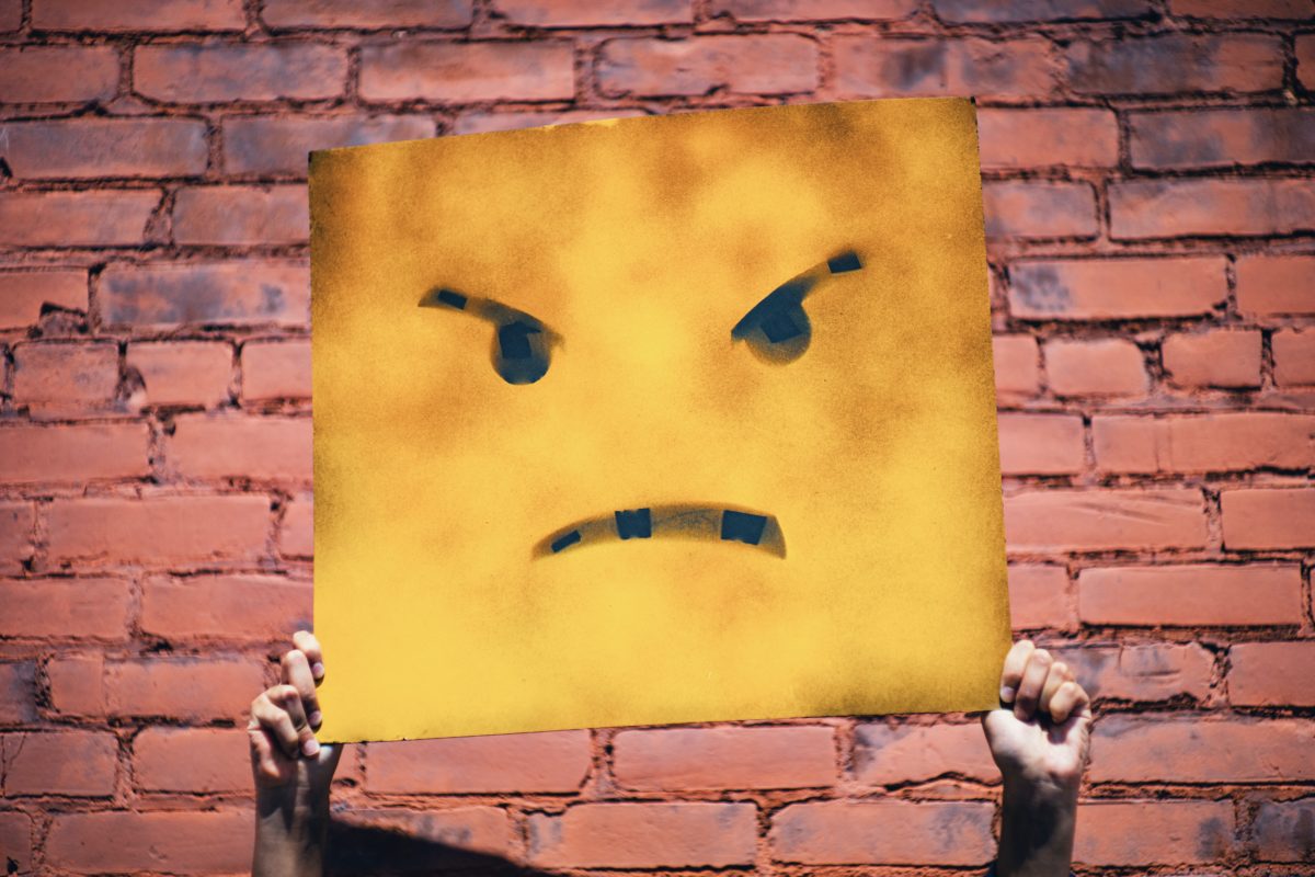 A pair of hands hold up a yellow sign that has an angry face on it. A brick wall is in the background.