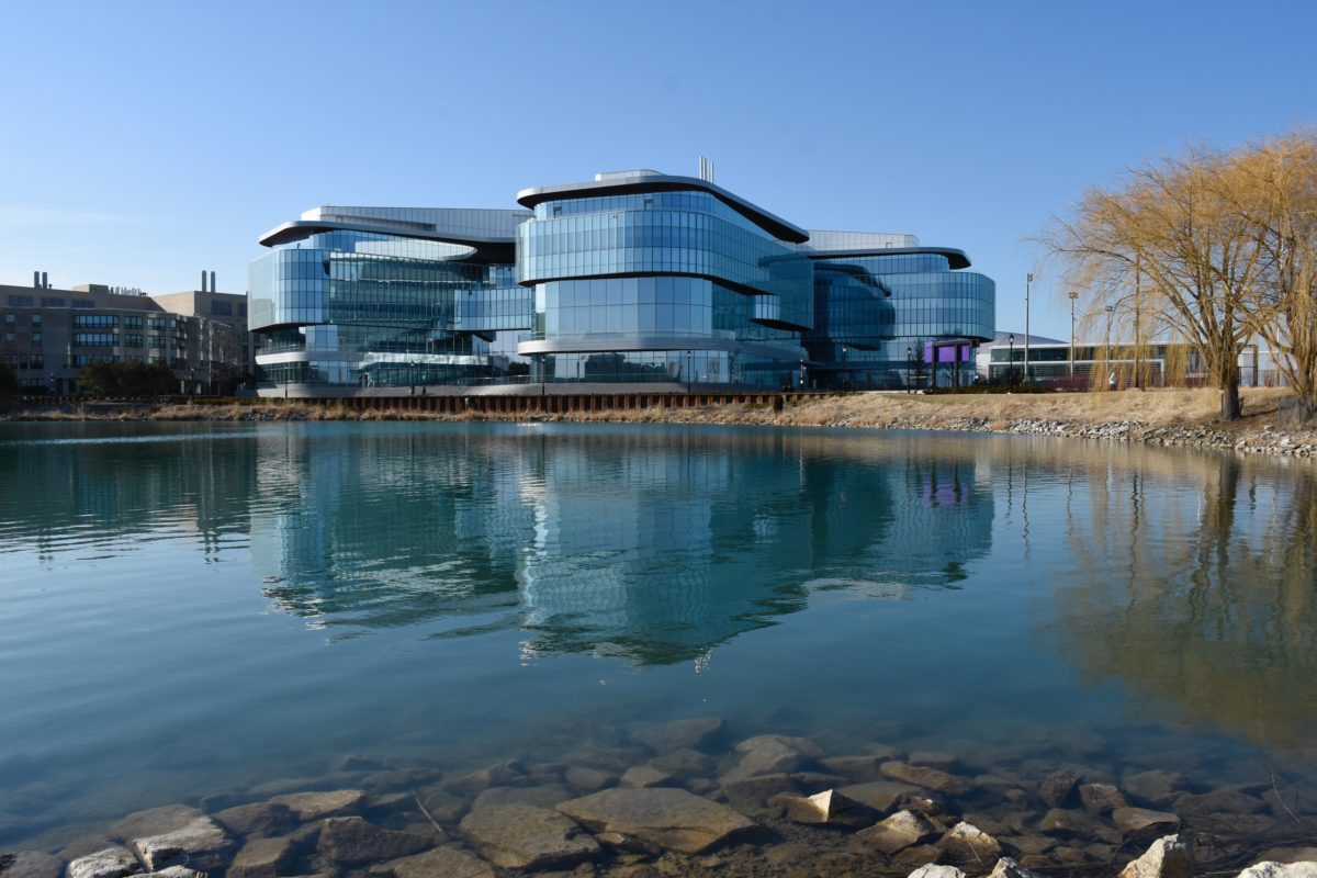 In the foreground is a small body of water. In the background is a large school building. The walls are all glass windows, and the building seems to have three main wings that jut out.