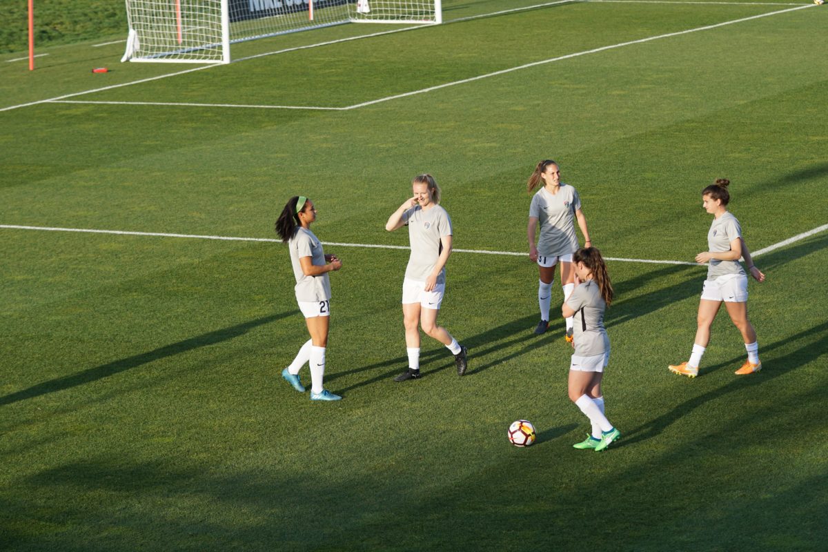 Five girls walk on a soccer field. They all wear white uniforms. A soccer ball lays at their feet.