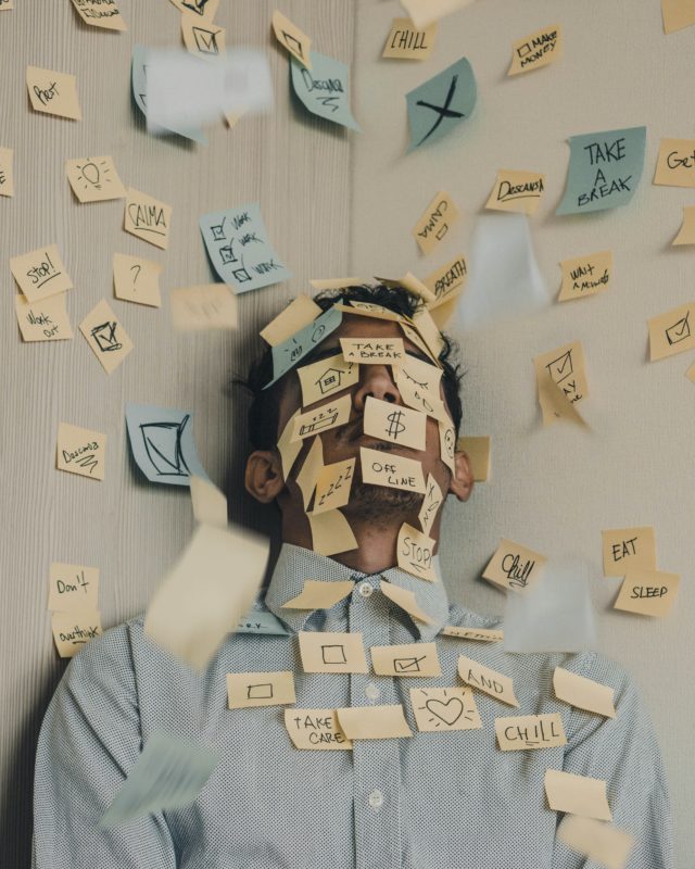 A man lays on a light wooden floor. He wears a button up shirt. His face and chest as well as the floor is covered in yellow and blue sticky notes. Some have small drawings and check marks while others have statements such as "stop!" or "chill"