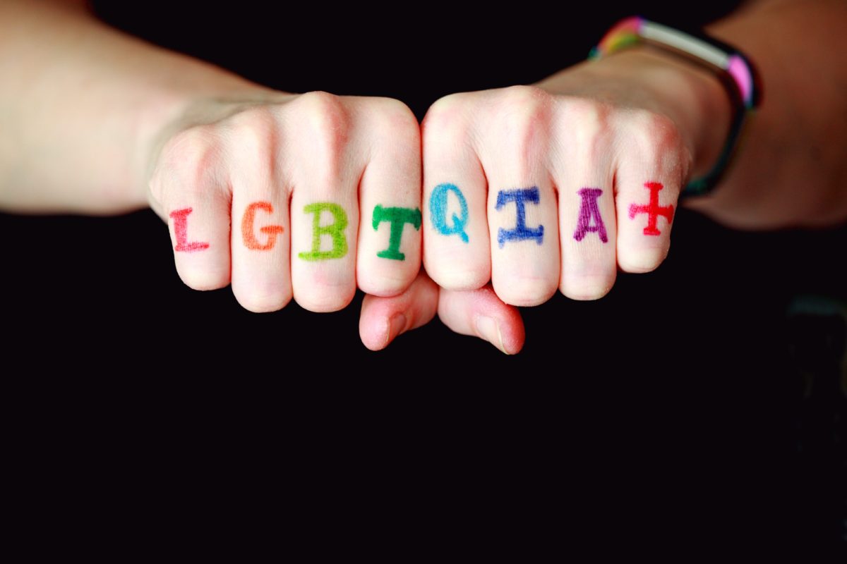 Two fists stick out toward the camera in front of a black background. On each knuckle is a letter. In order is says "LGBTQIA+" Each letter is a different color of the rainbow.