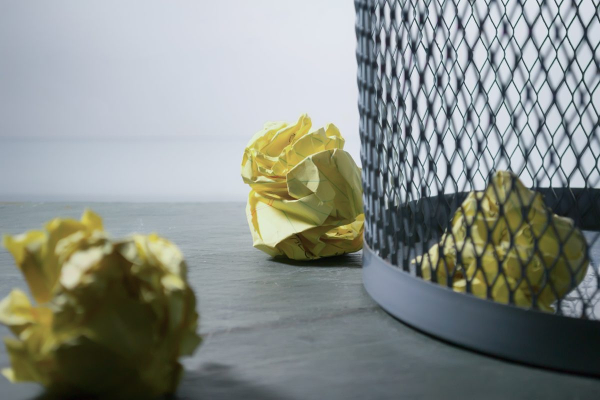 A small, metal trash can sits on a grey wood floor. Inside is a balled up piece of yellow paper. Outside of the can on the floor are two more pieces of balled up yellow paper.