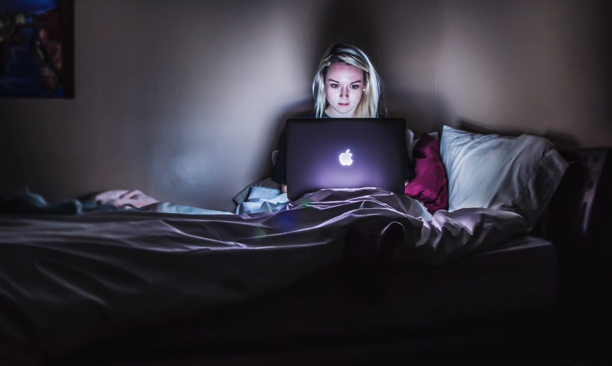 A white girl with short blond hair sits in her bed at night. The lights are out and she is illuminated by her Apple laptop. 
