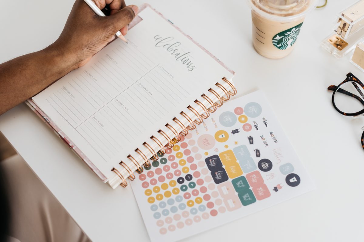 An overhead shot of someone writing in a planner. Next to the planner is a page of stickers and above it is a Starbucks coffee and a pair of glasses.
