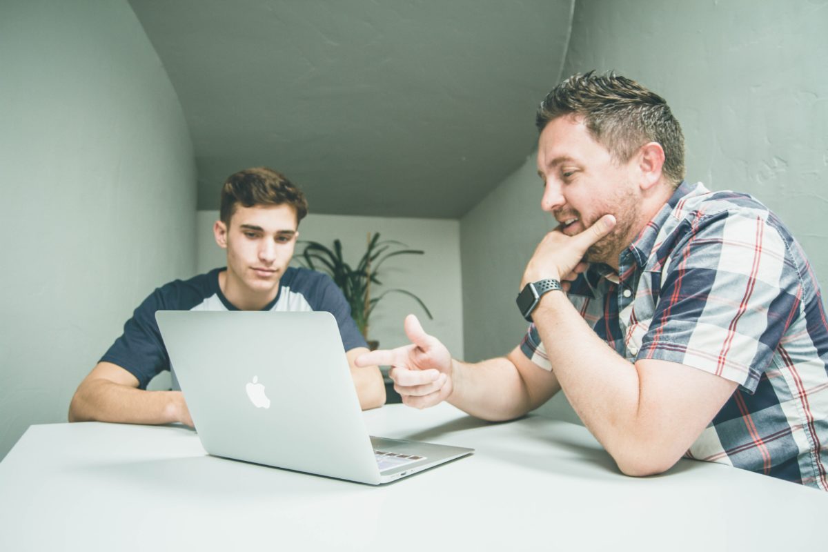 A white man with greying hair and a white teen boy with brown hair sit at a white table in a small room. The man points to his Apple laptop which sits on the table and they both look at the screen.