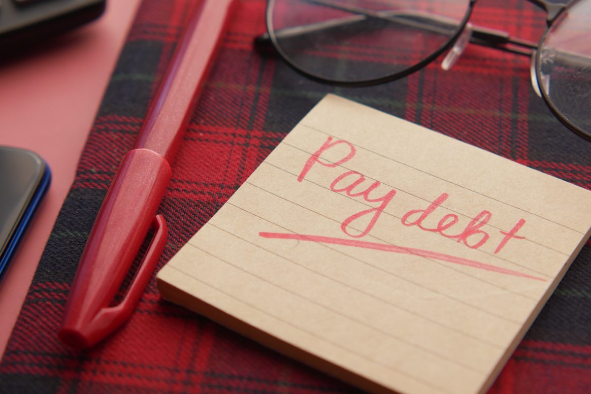 A yellow sticky note that says "Pay debt" in cursive red letters. The sticky note sits on a table covered in a red and black plaid cover. A red pen sits next to the sticky note and black glasses sit above it.