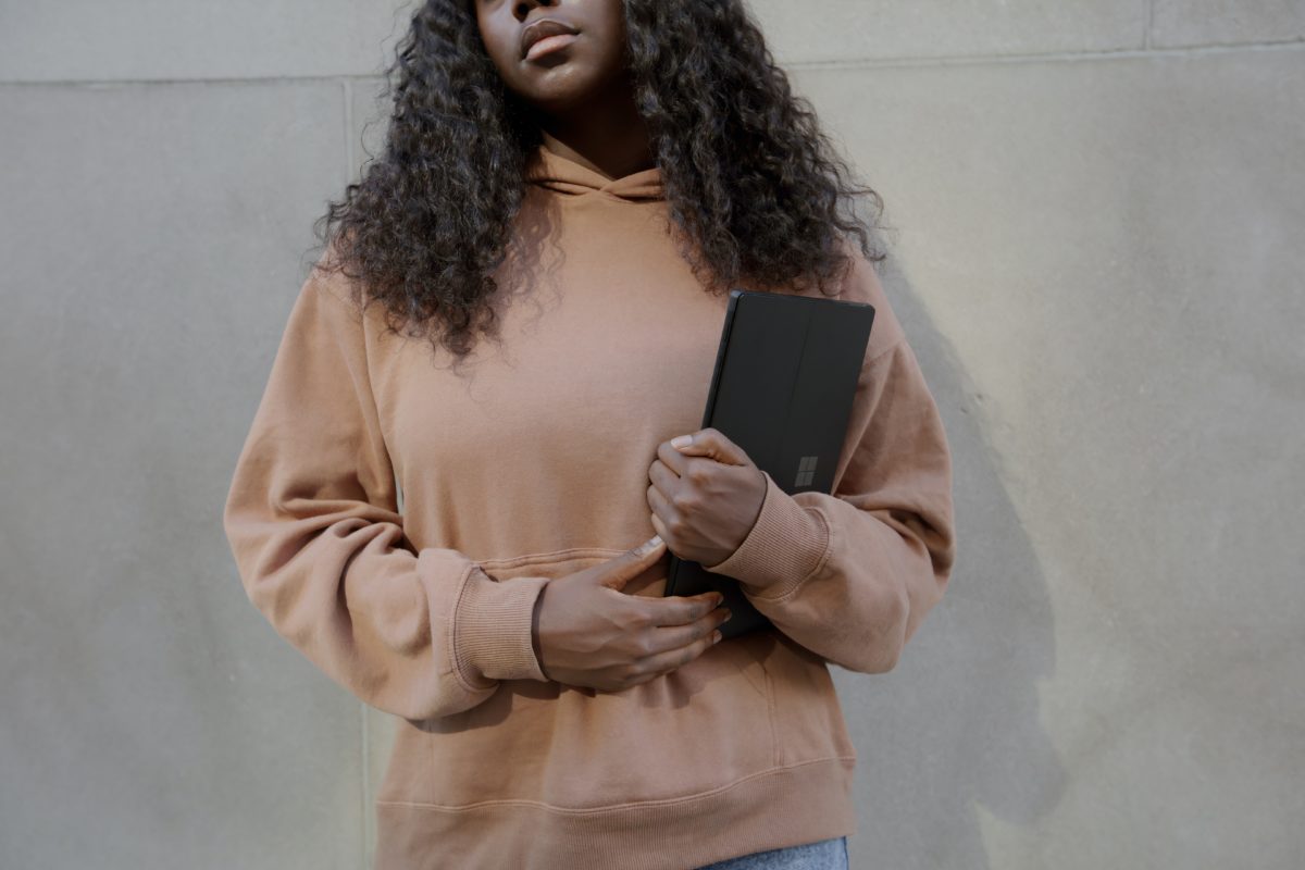 A young woman with dark brown skin and black curly hair leans against a grey stone wall. She wears a tan sweatshirt and holds a laptop in her arms.