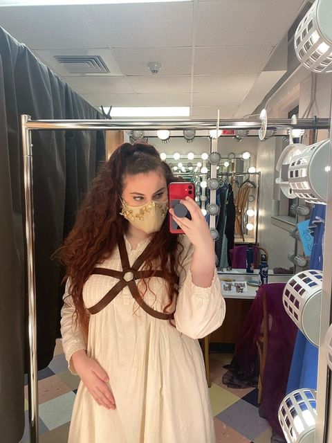 Anna, a young woman with light skin and long brown curly hair, takes a selfie in a mirror in a dressing room. Behind her is a black curtain, counters, and other mirrors with round lights. She holds her phone up near her face, looking at its screen out of the corner of her eye, and wears a tan mask, a long white flowy dress, and brown leather straps around her chest.
