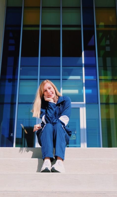 Erin, a young woman with light skin and blond hair, sits outside of a glass building on concrete steps. She rests her elbow on her knee and her head in her hand and smiles at the camera. She wears a blue track suit.