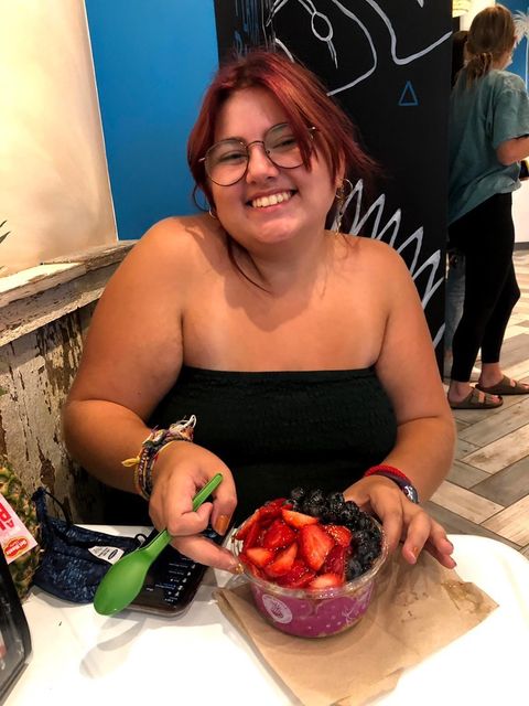 Amanda, a young woman with tan skin and brown/red hair, sits at a wooden table at a restaurant. She smiles at the camera and holds a fruit bowl in front of her. She wears a black tube top and black glasses.