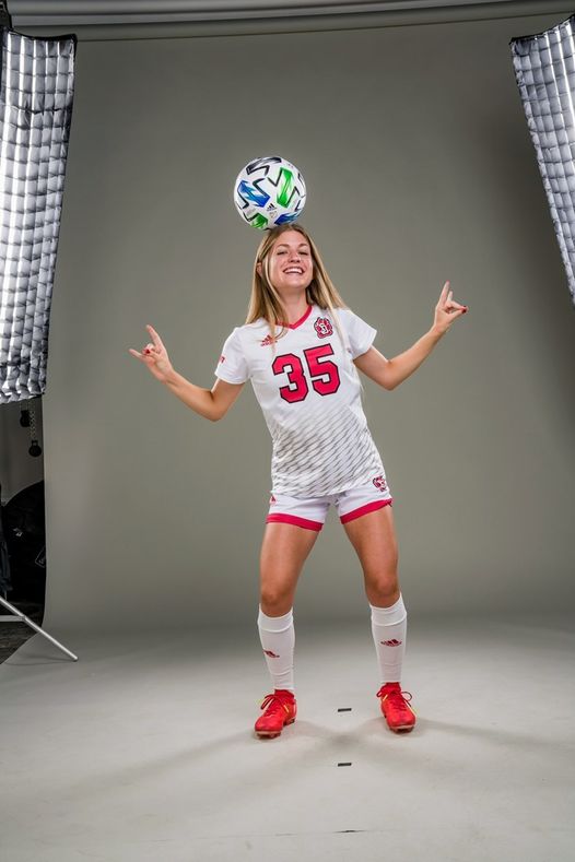 Kiah, a young woman with light skin and blond hair, stands in front of a grey backdrop. She bends her knees slightly and holds her hands up in horn signs. A soccer ball balances on her head and she wears a red and white soccer uniform.