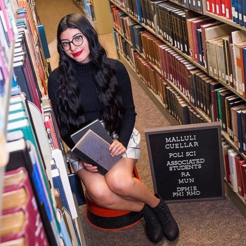 Malluli, a young woman with pale skin and long black hair, sits on a stool in between rows of books in a library. Next to her is a black sign that says "Malluli Cuellar Poli Sci Associated Students RA Mun PHIE PHI RHO". She holds two blue books in her lap and smiles at the camera while wearing a black and white skirt, black sweater, red lipstick, and black glasses.