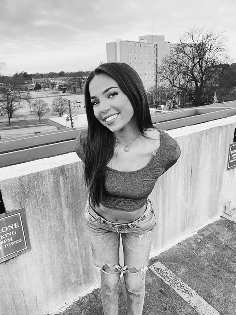 A black and white photo of Zoe, a young woman with tan skin and long black hair, stands outside at the top of a parking garage. Behind Zoe over the railing of the garage is a city street, trees, and a building in the distance. Zoe smiles at the camera and leans forward slightly, her arms behind her back. She wears jeans and a dark crop top.