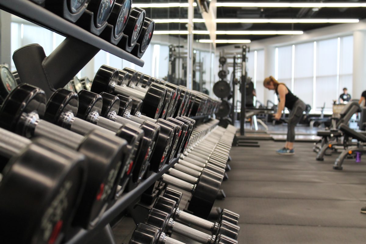 A rack of free weights in a gym. In the background is gym equipment and a woman working out.