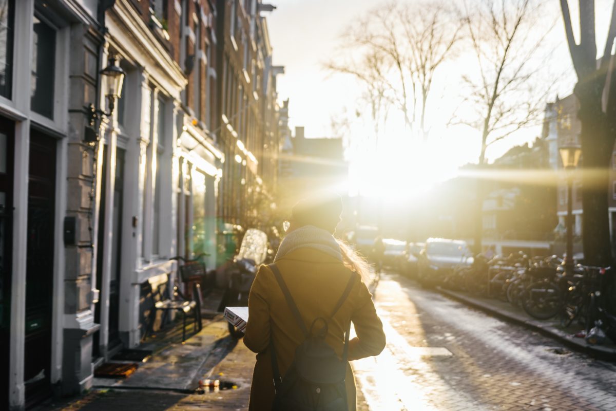 A woman walks down a city street, the sun shining brightly into the camera. From behind we see the woman holding a box and wearing a brown coat, grey scarf, black hat, and black backpack.