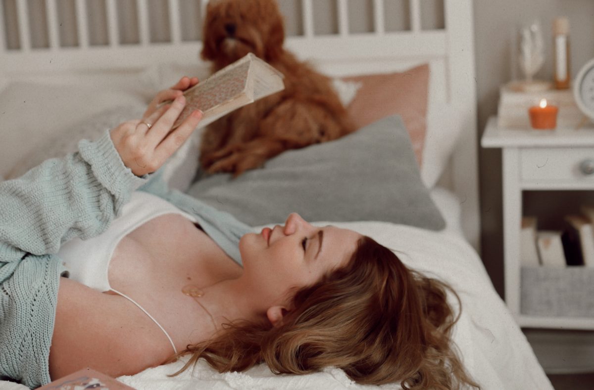 A young woman with light skin and brown hair lays horizontally across a bed with white covers. She wears a white tank top and a blue sweater and holds a book above her face that she reads. Behind her on the bed pillows is a small brown dog.