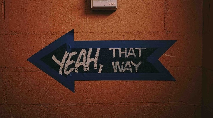 An orange concrete wall. Painted on it is a black arrow lined with blue tape. Written over the arrow in chalk it says "YEAH, THAT WAY"
