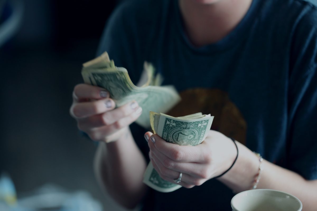 A person in a blue t-shirts sits and counts money, one dollar bills in each hand.