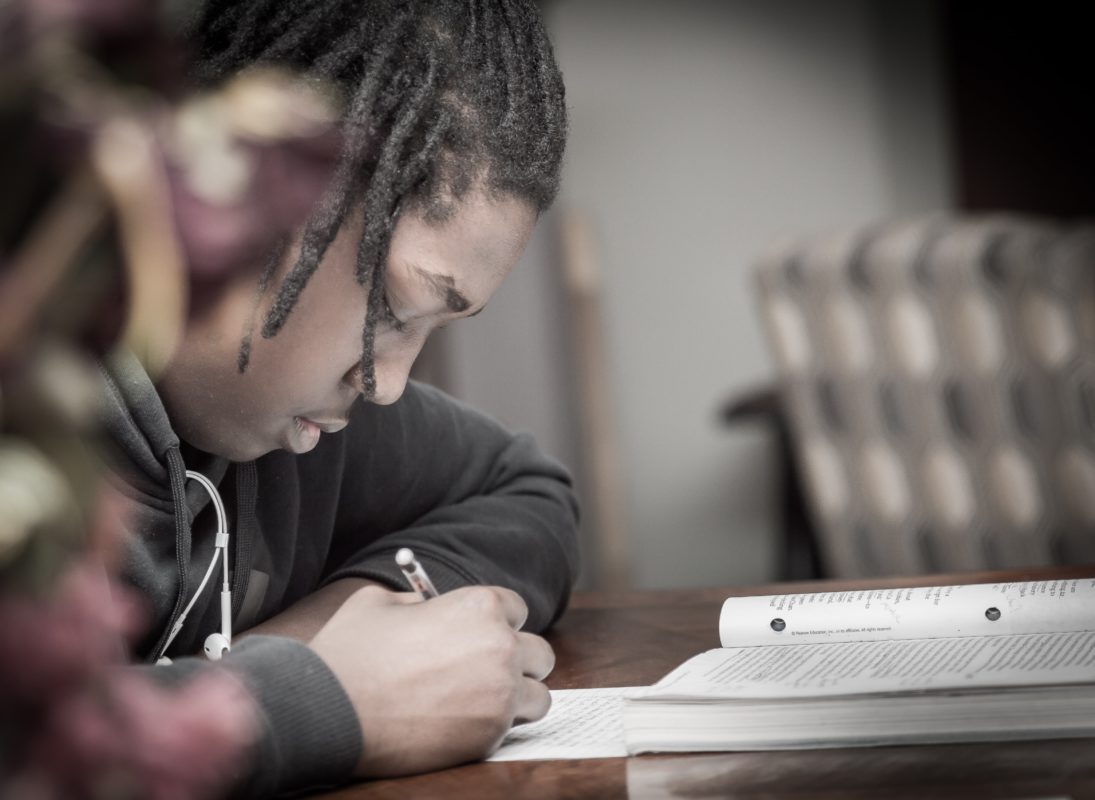 A young man with brown skin and locs sits at a desk. He leans down and puts his pencil to a piece of paper, his book sitting in front of him.