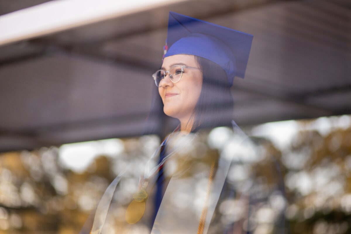A young woman with light skin and black hair in on the opposite side of a large window. She smiles and wears a blue graduation cap.