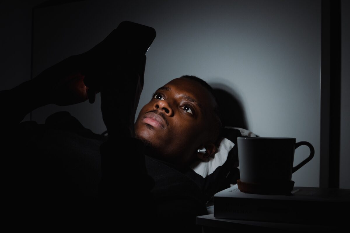 A young man with brown skin lays in bed at night. He holds his phone above him and it illuminates his face. To his side is a table with a book and coffee cup on it.
