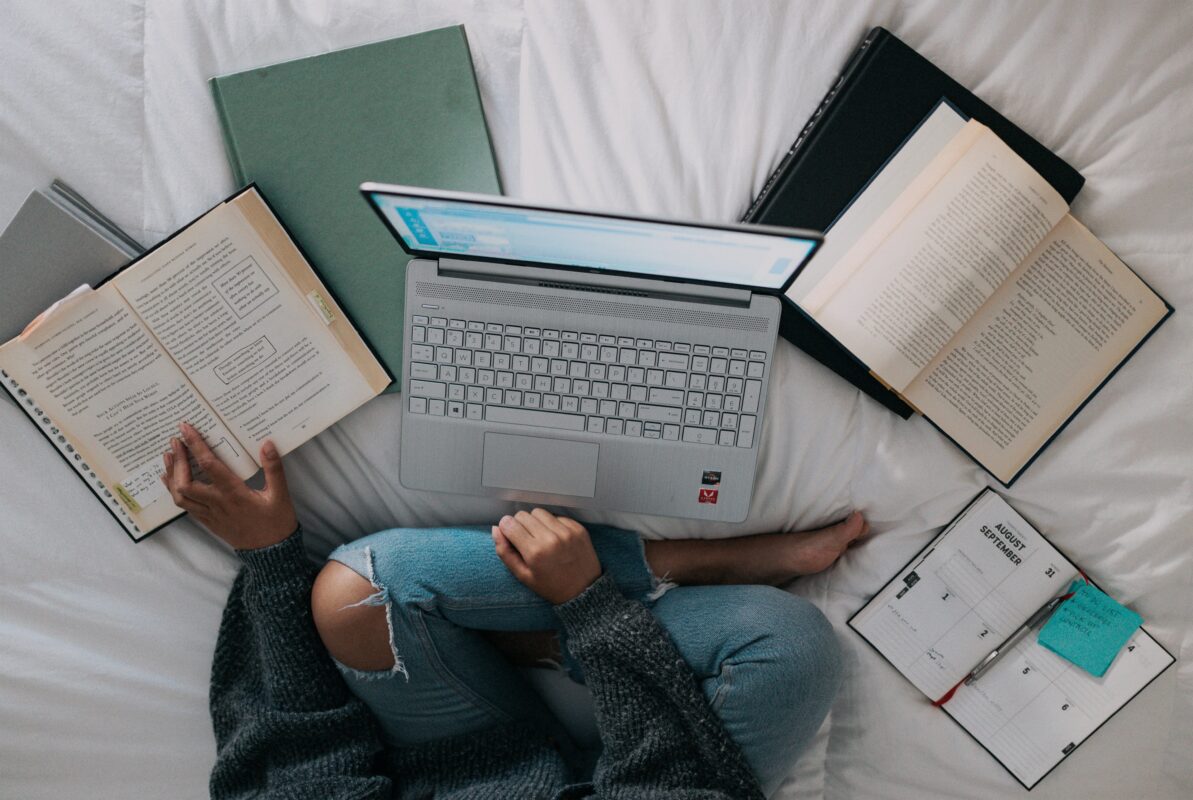 A person sits on a white bed with a computer and books splayed out in front of them.
