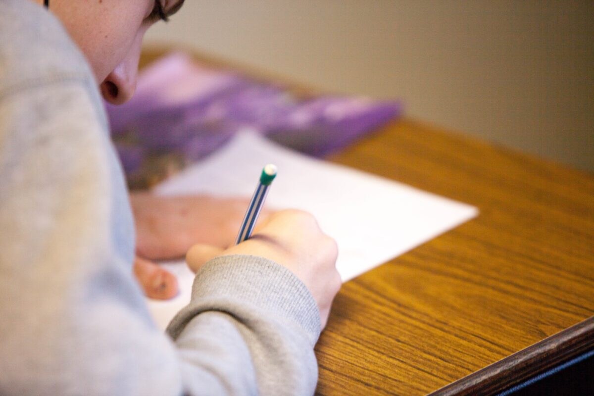 A young person with light skin leans over their desk and fills out a piece of paper with their pencil.