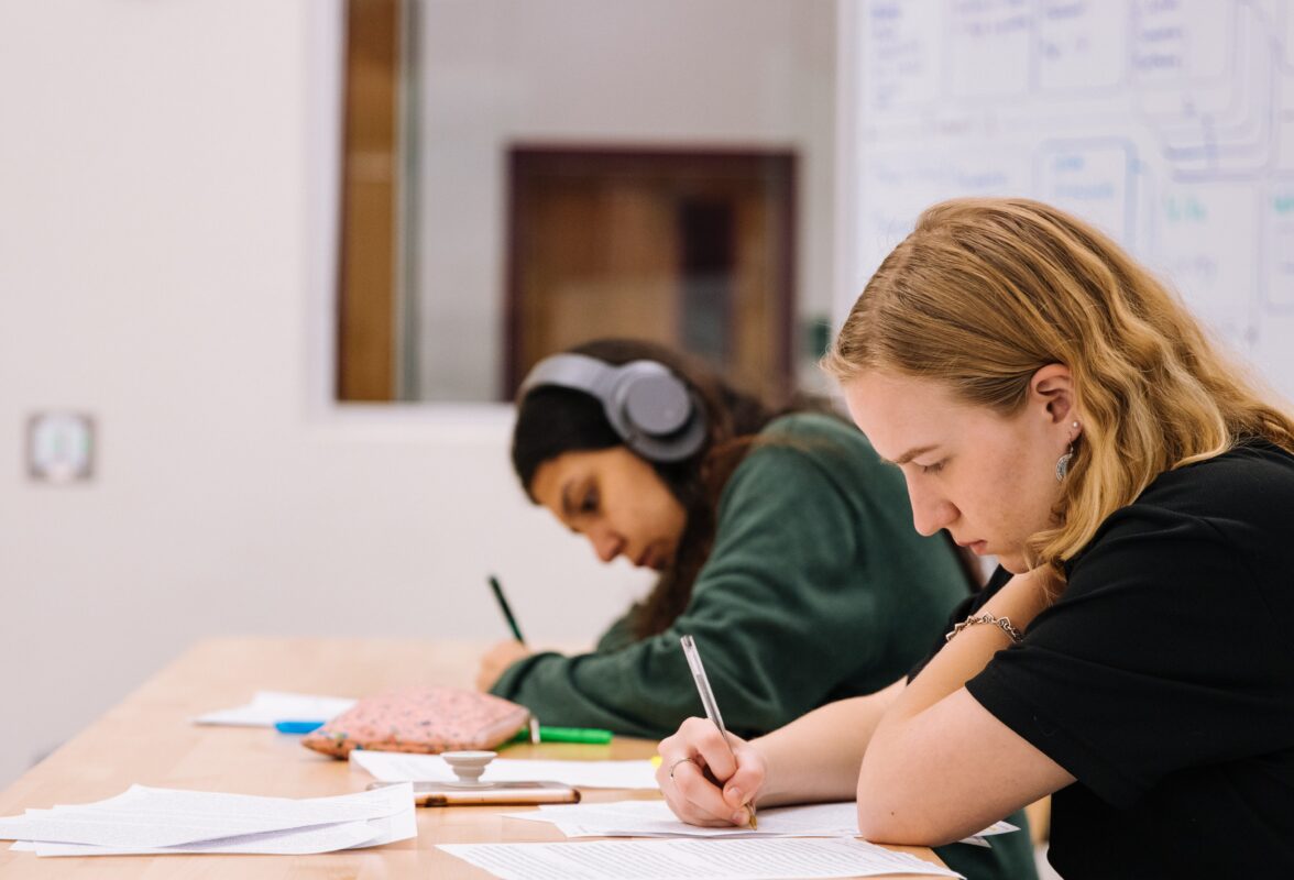 Two students sit at a desk in a classroom doing work.