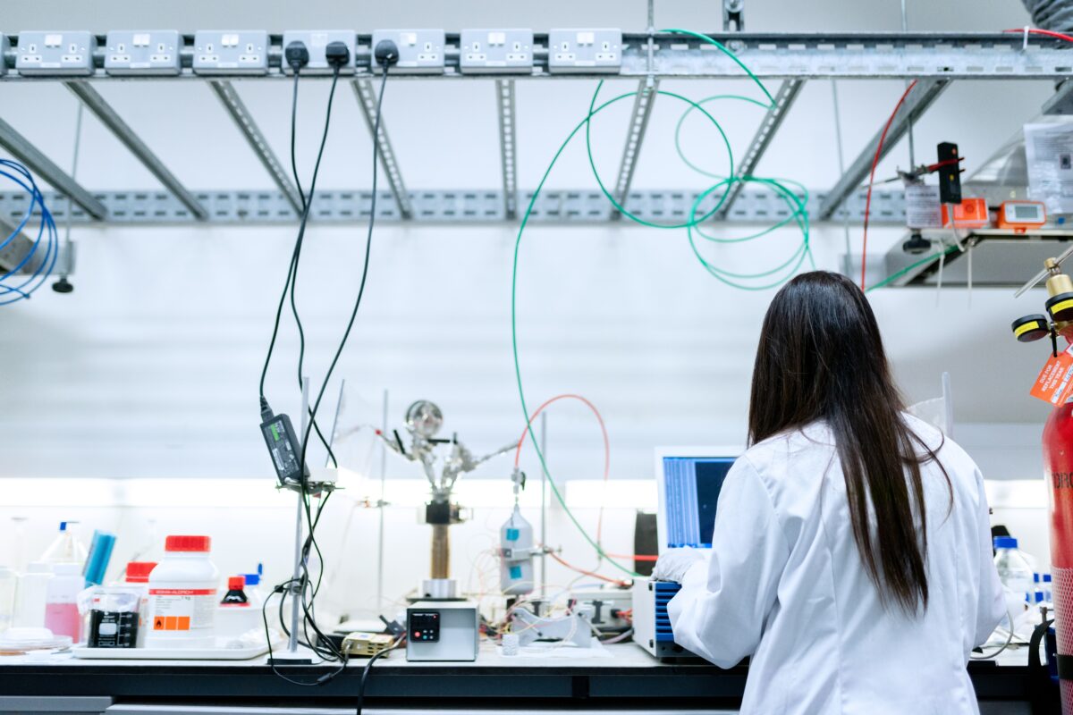 A woman stands in a lab with her back to the camera. The counter in front of her is covered in lab equipment.