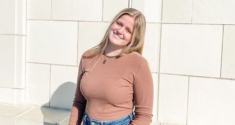 Leeya, a young woman with light skin and blond hair, stands in front of a white stone wall. She wears jeans and a brown shirt.