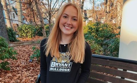 Skylar, a young woman with light skin and blond hair, sits on a bench in a park covered in orange leaves. She displays her black Bryant University sweatshirt.
