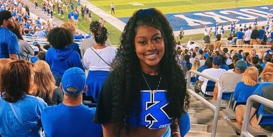 Natya, a young woman with brown skin and brown curly hair, stands in the stands of a football stadium. Behind her is the field and seated fans. She wears blue KY merch.