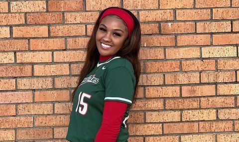 Jaylon, a young woman with brown skin and brown hair, poses in front of a brick wall. She smiles over her shoulder and wears a red and green softball uniform.