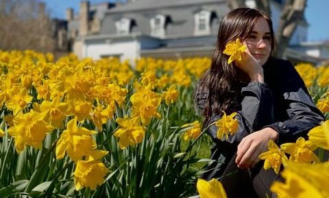 Kaleigh, a young woman with light skin and brown hair, sits in a field of yellow daffodils. She rests her head in her hand and looks off to the side.