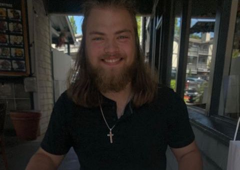 Luke (a young man with light skin, long brown hair, and a beard) sits at a table outside, a sandwich sitting in front of him. He wears a black tee.