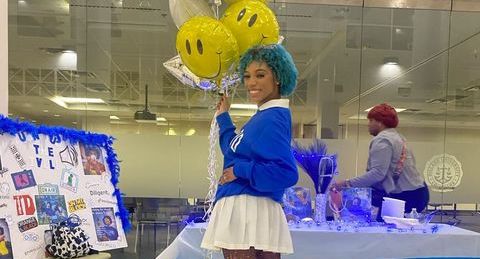 Kevell (a young woman with brown skin and blue hair) stands in front of a blue table holding a bunch of yellow smiley face balloons.