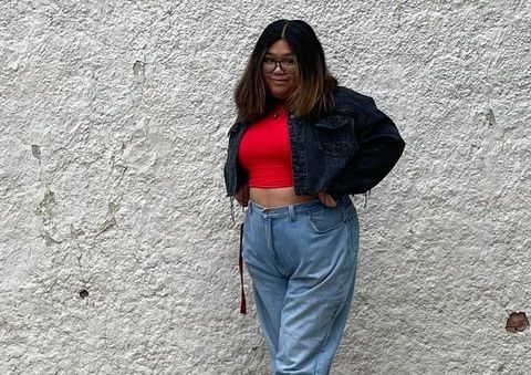 Samantha (a young woman with tan skin and brown hair) poses in front of a white stone wall. She wears jeans, a red crop top, and blue jacket.