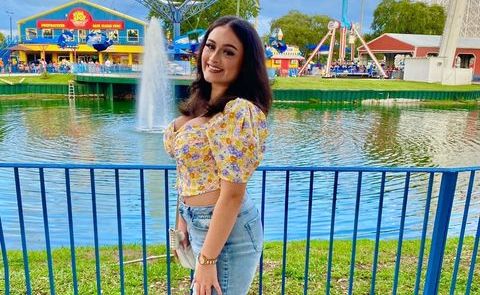 May (a young woman with tan skin and brown hair) poses in front of a view of a pond and carnival rides. She wears jeans and a yellow floral blouse.