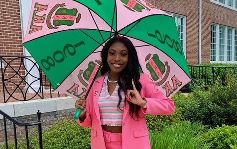 Kayla (a young woman with brown skin and brown hair) poses outside on the sidewalk. She wears a bright pink suit and holds a pink and green Alpha Kappa Alpha umbrella. She holds up her hand and makes an Alpha Kappa Alpha symbol.