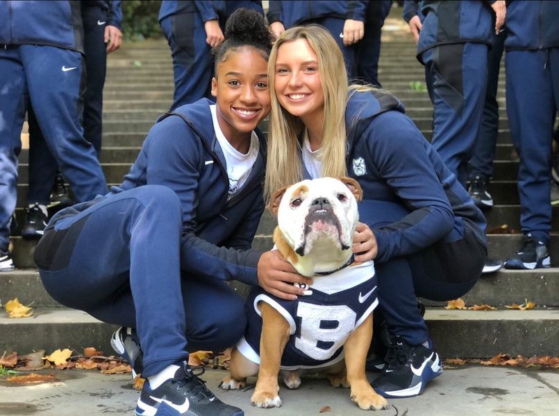 Alani (a young woman with brown skin and brown curly hair) kneels down next to a bulldog in a Butler jersey with a friend (a young woman with light skin and blond hair)