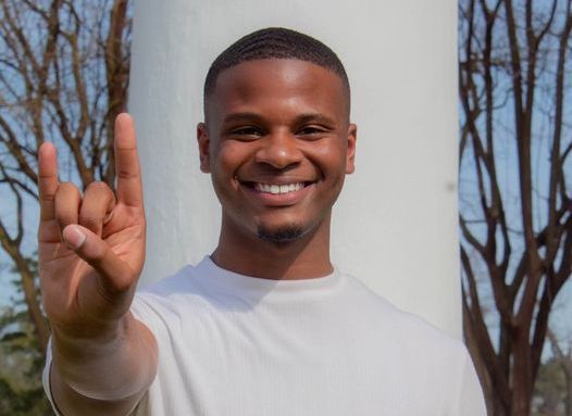 TJ (a young man with brown skin and short brown hair) stands outside in front of a large pillar. He holds out a "fork em" sign with his hand in front of him.
