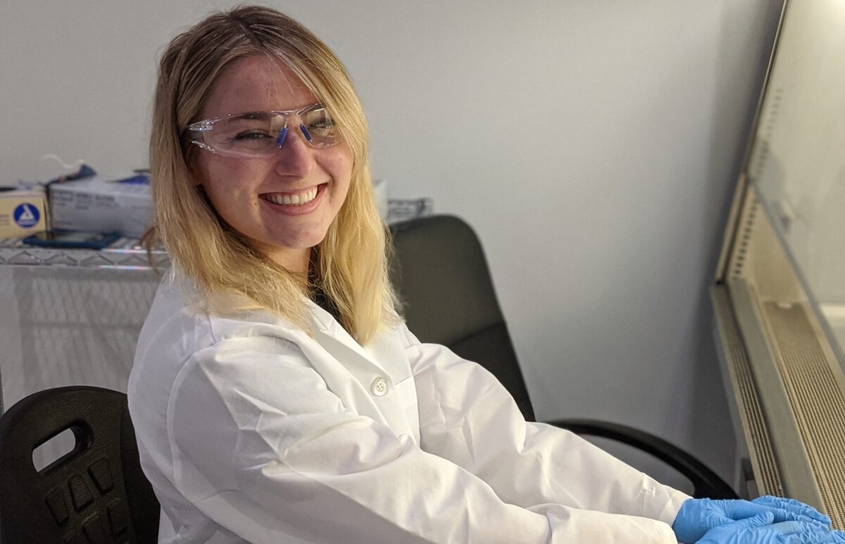 A young woman with light skin and blond hair sits at a desk in a lab wearing safety glasses, a lab coat, and blue gloves.