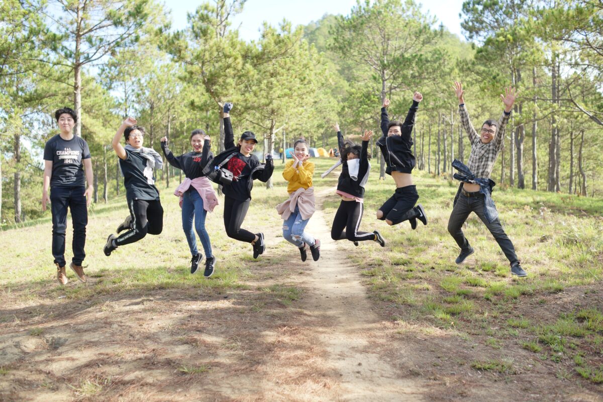 A group of students jumps on a trail in the woods.