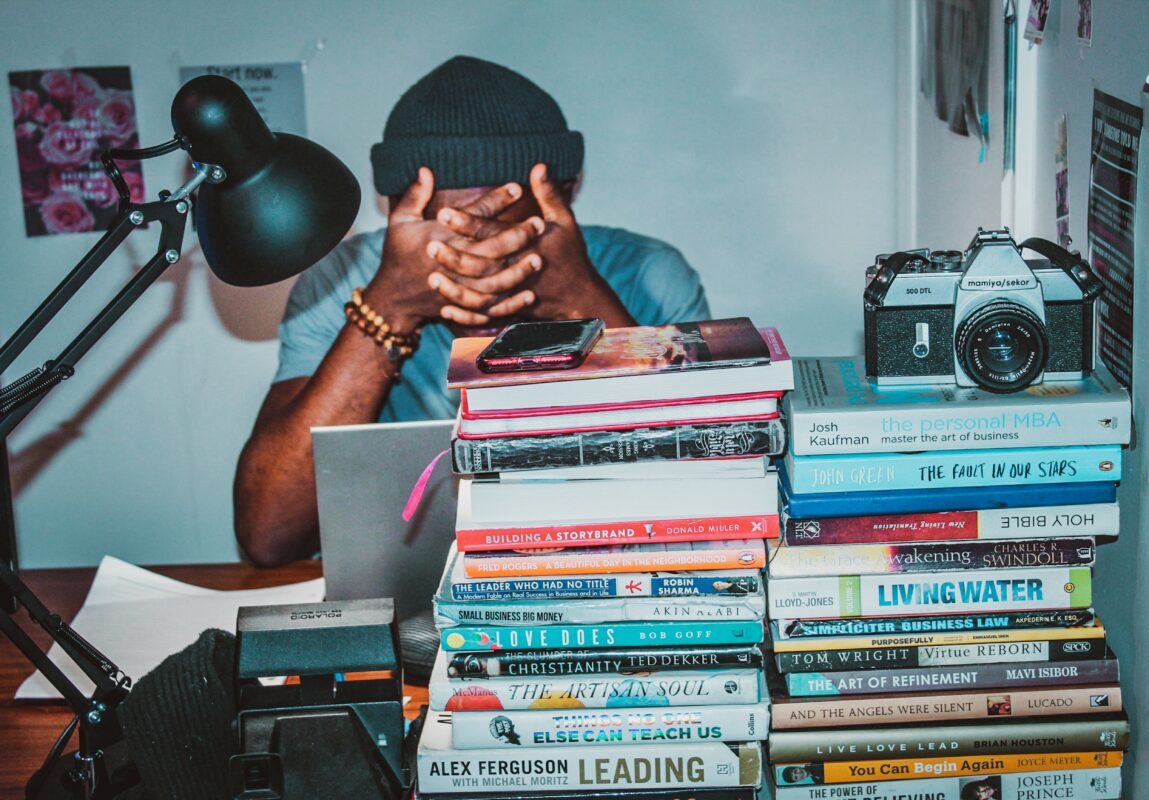 A young man with brown skin sits at a desk. In front of him are tall stacks of books. He puts his face in his hands in frustration.