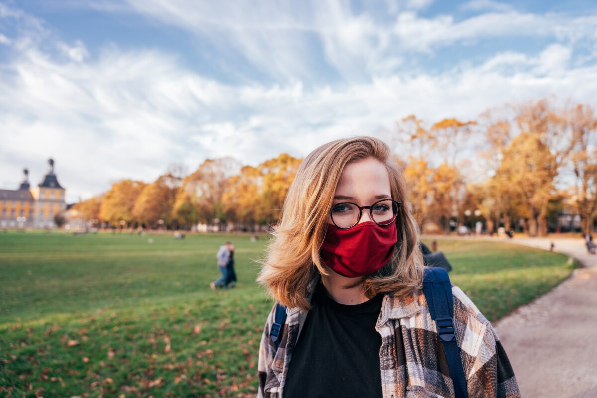 A young woman with light skin and blond hair stands on a college campus wearing a bright red mask.